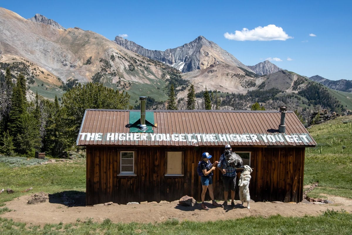 Couple with two dogs standing in front of Pioneer Cabin in Sun Valley. On the roof it says "the higher you get, the higher you get"